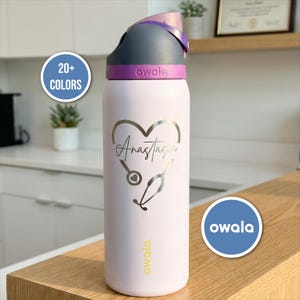 May include: A light pink Owala water bottle with a gray and purple lid. The bottle features a heart design with the name "Anastasia" and a stethoscope. The text "20+ COLORS" and "owala" are also visible.