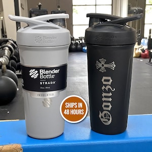 May include: Two Blender Bottle insulated shaker bottles. One is light gray with a black label that reads "STRADA" and holds 24 oz. The other is black with a cross design and the word "Gonzo". Both have flip-top lids.