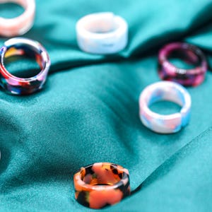 May include: A selection of resin rings in diverse colours and patterns. The rings showcase shades of pink, blue, orange, and purple, with some exhibiting a marbled appearance. They are presented on a teal satin fabric.