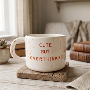 May include: A white ceramic mug with the words "CUTE BUT OVERTHINKER" in red lettering. The mug has a large handle and is speckled with red paint. It sits on a small, square wooden coaster.