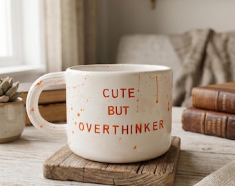 May include: A cream-colored ceramic mug with a rounded handle and orange paint splatters. The text "CUTE BUT OVERTHINKER" is printed in orange on the front. The mug has a smooth finish and a wide, cylindrical shape with a flat base.