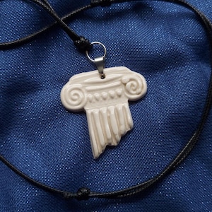 May include: A cream-colored pendant shaped like a broken Ionic column capital with a smooth, glossy finish. The pendant is attached to a thin, black cord necklace by a silver-toned metal jump ring and a small metal bail at the top center.