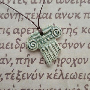 May include: A ceramic pendant necklace featuring a stylized, light green and white Ionic column design. The pendant is suspended from a brown cord, set against a backdrop of faded text.