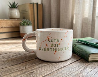 May include: A white ceramic mug with a rounded handle, featuring orange paint splatters and the green text "CUTE BUT OVERTHINKER" printed on the front. The mug has a smooth, glossy finish and a wide, low-profile cylindrical shape.