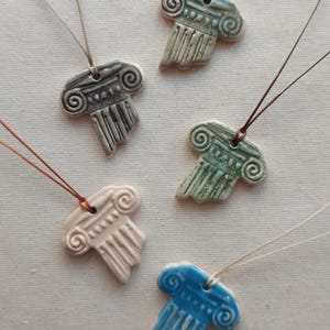 Puede incluir: Una colección de collares con colgantes de cerámica con forma de columna en varios colores, incluyendo gris, blanco, verde azulado y azul. Cada colgante está suspendido de un cordón fino de color neutro, mostrando un diseño arquitectónico clásico.
