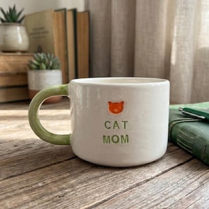 May include: A white ceramic mug with a light green handle, featuring an orange cat head icon above the green text "CAT MOM" embossed on the front. The mug has a smooth, rounded shape and a glossy finish, showcasing a simple and clean design.