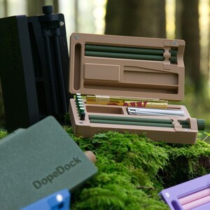 Könnte beinhalten: Eine Auswahl an Etuis und Zubehör, darunter ein schwarzes vertikales Etui, ein beiges Etui mit grünen und silbernen Komponenten und ein grünes Etui mit dem Text "DopeDock". Die Artikel werden auf grünem Moos präsentiert.