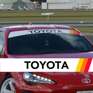 Op de afbeelding: Rode Toyota sportwagen met een witte banner over de voorruit. De banner toont het Toyota-logo in zwarte tekst. Een kleurrijk streepontwerp in geel, oranje en rood accentueert de rechterkant van de banner.