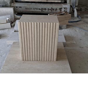 May include: A beige, rectangular stone block with vertical grooved detailing on one side. The block is sitting on a larger, matching stone slab. The background includes machinery and other stone pieces, suggesting a construction or manufacturing setting.