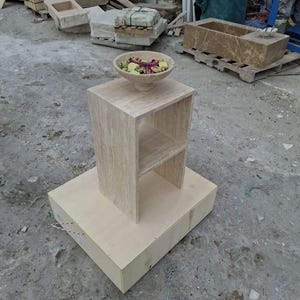 Puede incluir: Una pequeña mesa auxiliar de madera marrón claro con un estante. La mesa está sentada sobre una base de madera marrón claro más grande. Un pequeño tazón lleno de flores moradas y verdes se sienta encima de la mesa.