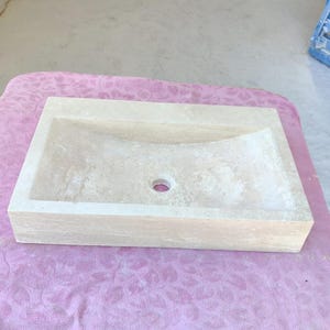 May include: A rectangular, beige stone sink with a gently curved basin and a central drain hole. The sink has a textured surface and a simple, modern design, suitable for bathroom or powder room use.