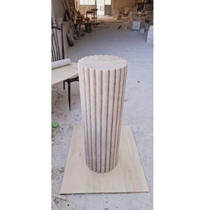 May include: A tall, cylindrical pedestal with vertical fluting, crafted from light-colored stone. The top and bottom are flat, and the pedestal stands on a wooden base. The fluted design adds texture and visual interest to the sculpture.
