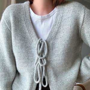 May include: A light gray knitted cardigan with a V-neck and a tie detail at the front. The cardigan is made of a soft, comfortable material and has a relaxed fit.
