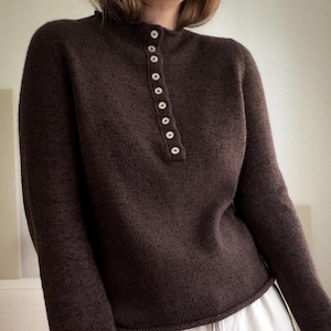 May include: A dark brown long-sleeved jumper featuring a high neckline and a vertical placket down the centre front. The placket is secured with seven small, round, light-coloured buttons. The jumper has a simple knit texture and long sleeves.