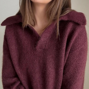 Może przedstawiać: Bordowy sweter z dzianiny z dekoltem w kształcie litery V i kołnierzem. Sweter wykonany jest z miękkiego, puszystego materiału.