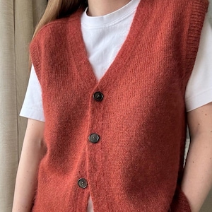 Op de afbeelding: Een roestkleurige gebreide vest met een V-hals en knoopsluiting. De vest is gemaakt van een zacht, pluche materiaal en heeft een ontspannen pasvorm.