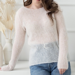 以下が含まれることがあります： ゆったりとしたシルエットと長袖の白い軽量ニットセーター。柔らかく繊細な素材で作られており、わずかに透き通った外観をしています。