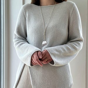 Puede incluir: Un suéter de punto gris claro con un dobladillo asimétrico único. La parte superior de manga larga presenta un cuello barco y se combina con un collar largo plateado con un colgante. El suéter es de color neutro, adecuado para varios atuendos.