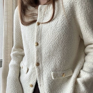 May include: A white cardigan with a textured knit and gold buttons. The cardigan has two pockets on the front and a slightly cropped length.