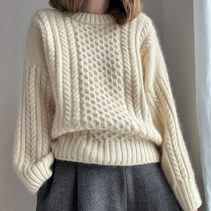 May include: Cream-colored cable knit sweater with a round neckline and ribbed cuffs. The sweater features a textured pattern on the front and sleeves. The garment is paired with a gray skirt.