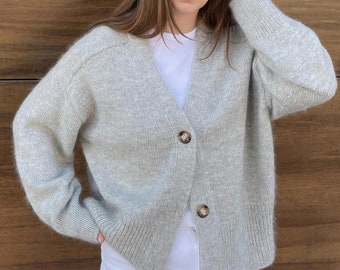 May include: A light gray knitted cardigan with a V-neck and button closure. The cardigan has long sleeves and a relaxed fit.