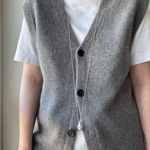 Op de afbeelding: Een grijs gebreid vest met een zilveren kettingketting en een zilveren hanger. Het vest heeft een V-hals en een knoopsluiting.