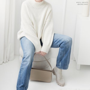 Pu&ograve; includere: Una donna indossa un maglione bianco a collo alto e jeans blu e siede su uno sgabello tenendo una borsa a mano marrone chiaro. #NESS_SWEATER @MORECAKNIT