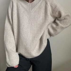 May include: A cream-colored knitted sweater with a v-neck and long sleeves. The sweater is made of a soft, textured yarn and has a relaxed fit.