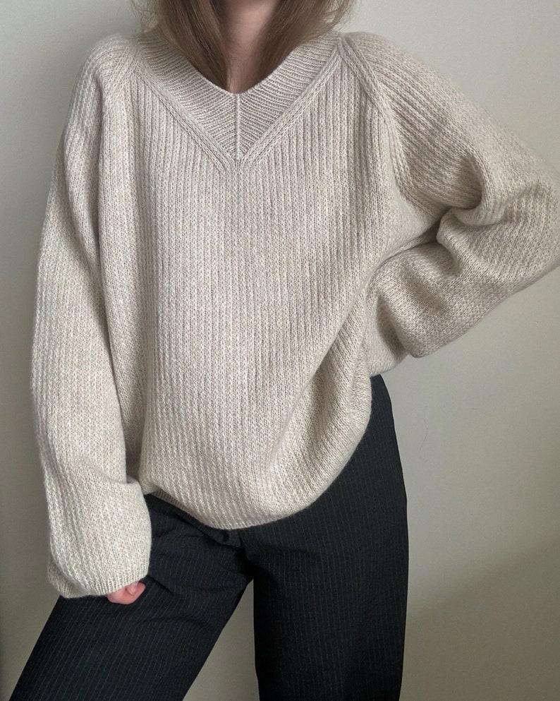 May include: A cream-colored knitted sweater with a v-neck and long sleeves. The sweater is made of a soft, textured yarn and has a relaxed fit.