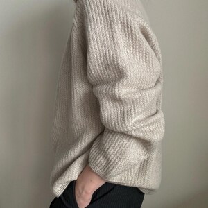 May include: A cream-colored knitted sweater with a ribbed texture. The sweater has long sleeves and a relaxed fit.