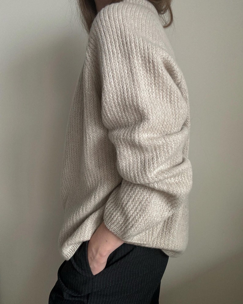 May include: A cream-colored knitted sweater with a ribbed texture. The sweater has long sleeves and a relaxed fit.