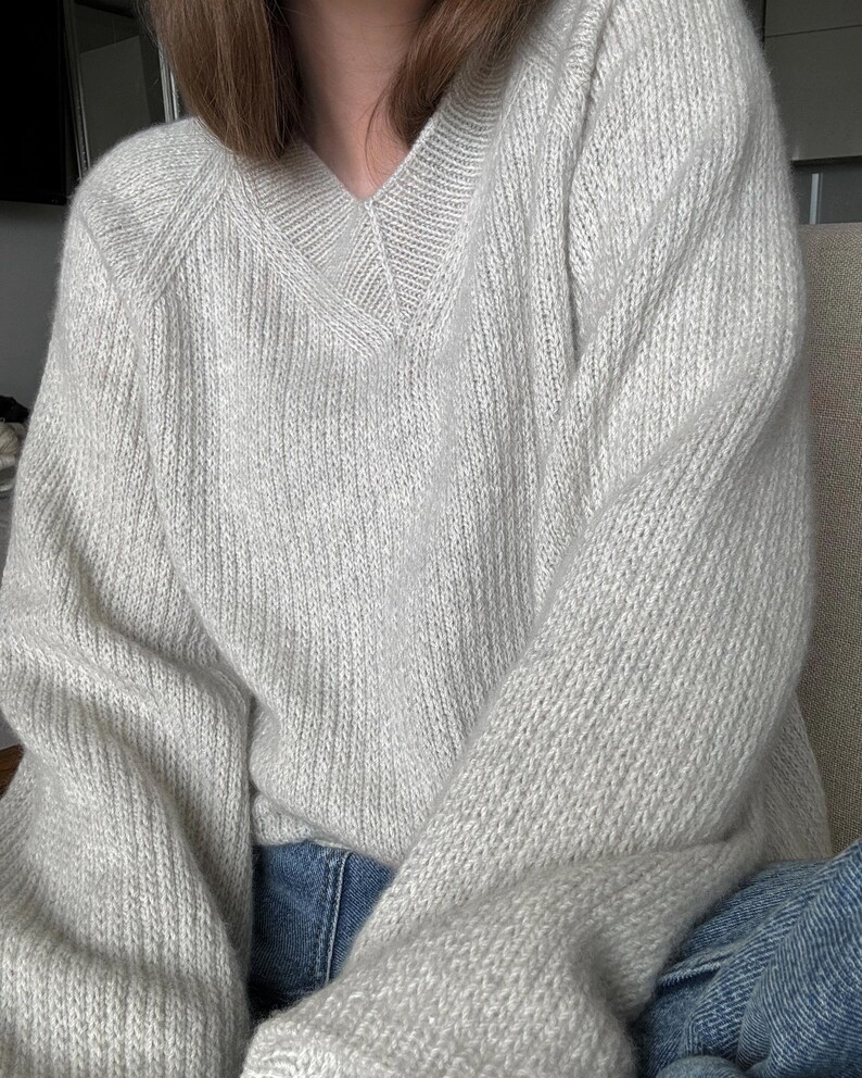 May include: A light gray knitted sweater with a v-neck and long sleeves. The sweater is made of a soft, fuzzy material.