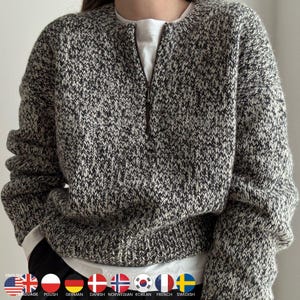 May include: A speckled grey and white knit sweater with a zip-up front. The sweater has a round neckline and long sleeves. A white t-shirt is visible underneath. The sweater is a casual style.