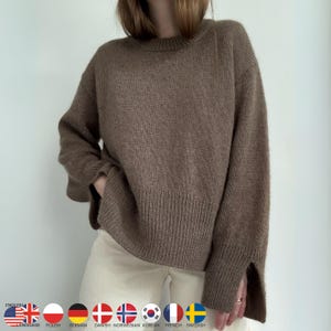 Peut inclure: Un pull brun surdimensionné avec un ourlet et des poignets côtelés. Le pull a un col rond et des manches longues avec une fente au poignet. Le modèle porte le pull avec un pantalon crème.