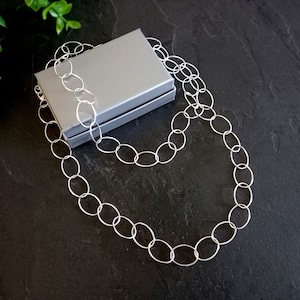 May include: A silver necklace with oval links is draped over a grey jewellery box. The necklace is displayed on a dark, textured surface. The links are uniform in size and shape, creating a simple, elegant design.