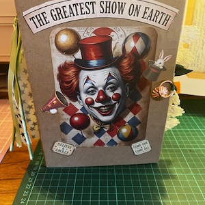 Puede incluir: Una cubierta de álbum de recortes color canela con el texto THE GREATEST SHOW ON EARTH, una ilustración central de un payaso con sombrero de copa rojo, un conejo en un sombrero, globos y un megáfono. Dos etiquetas pequeñas dicen BELIEVE & AMAZE y COME ONE COME ALL.