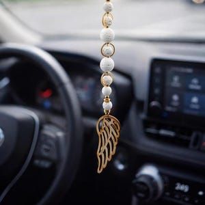 Könnte beinhalten: Ein Auto-Charm mit einem goldfarbenen Flügelanhänger und einer Kette aus weißen, strukturierten Perlen. Der Charm hängt am Rückspiegel. Ein kleines herzförmiges Etikett trägt die Aufschrift "HAND MADE WITH LOVE."
