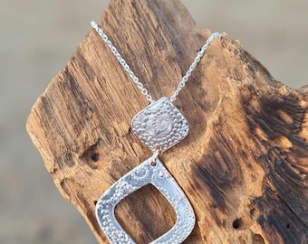 May include: A silver necklace with two geometric pendant charms. The charms are textured with a repeating pattern of small circles. The necklace is displayed on a piece of driftwood.