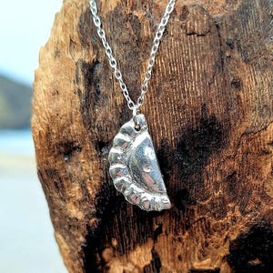 May include: A silver necklace with a charm shaped like a half-eaten pastry. The charm has a textured crust and a smooth filling. The necklace is displayed on a piece of driftwood.