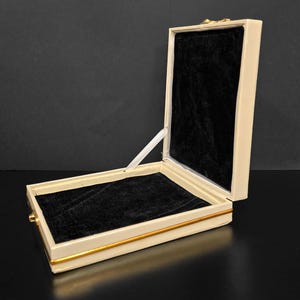 Puede incluir: Una caja de joyería blanca con un borde dorado y un interior de terciopelo negro. La caja está abierta y la tapa está levantada.