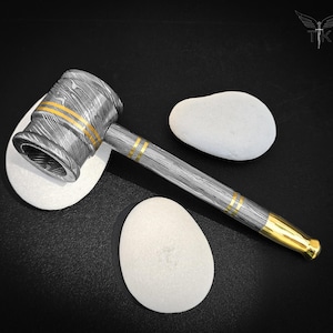 May include: A detailed, Damascus steel hammer with gold accents. The hammer head and handle are intricately patterned with a layered steel design. The handle features gold bands, and the end is capped with gold. The hammer rests on a black surface with white stones.