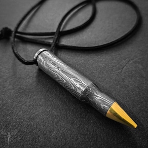 Könnte beinhalten: Eine Halskette mit einem silbernen Patronen-Anhänger mit goldener Spitze, aus gemustertem Metall, an einem schwarzen Lederband.