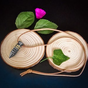 Könnte beinhalten: Ein silberner und goldener Anhänger in Form einer Kugel mit einer Lederschnur auf zwei Holzkreisen mit grünen Blättern.