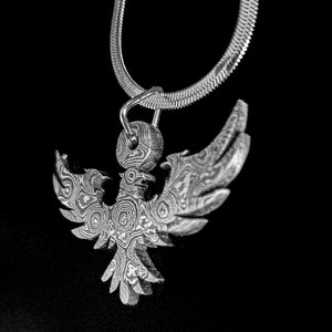 Pode incluir: Um colar de prata com um pingente de fênix detalhado e estampado. O pingente é feito de metal com um design de redemoinho e textura.