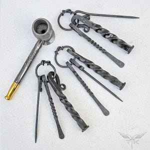 Op de afbeelding: Een handgesmede metalen pijp en diverse pijpgereedschappen. De pijp heeft een patroonafwerking met een goudkleurige punt. De gereedschappen omvatten gedraaide en rechte metalen stukken die aan ringen zijn bevestigd. De items zijn op een lichtgekleurd oppervlak geplaatst.