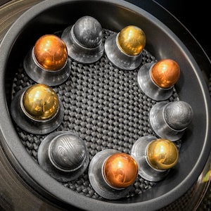 May include: An overhead shot of eight metal objects arranged in a circle. Each object has a silver base and a spherical top in either gold, copper, or silver. The objects are set in a dark, textured container.