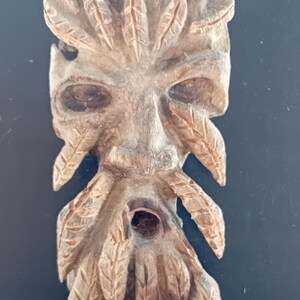 May include: A wooden carving of a green man's face with leaves for hair and beard. The carving is detailed and realistic, with a hole in the mouth.