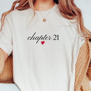 Peut inclure: T-shirt crème avec l'inscription "chapter 21" en écriture cursive, un petit cœur rouge en dessous. Le t-shirt fait partie d'une tenue décontractée, porté avec un cardigan beige.