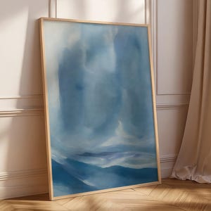 May include: A framed abstract art print featuring a watercolor painting in shades of blue. The artwork depicts a landscape with soft, blurred forms suggesting mountains and clouds. The frame is a light wood color.