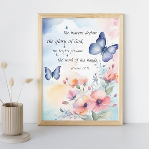Puede incluir: Una impresión en acuarela enmarcada con el texto "Los cielos proclaman la gloria de Dios", ilustraciones florales y de mariposas. La obra de arte tiene un marco de madera clara y una paleta de colores pastel suaves de azules, rosas y amarillos.